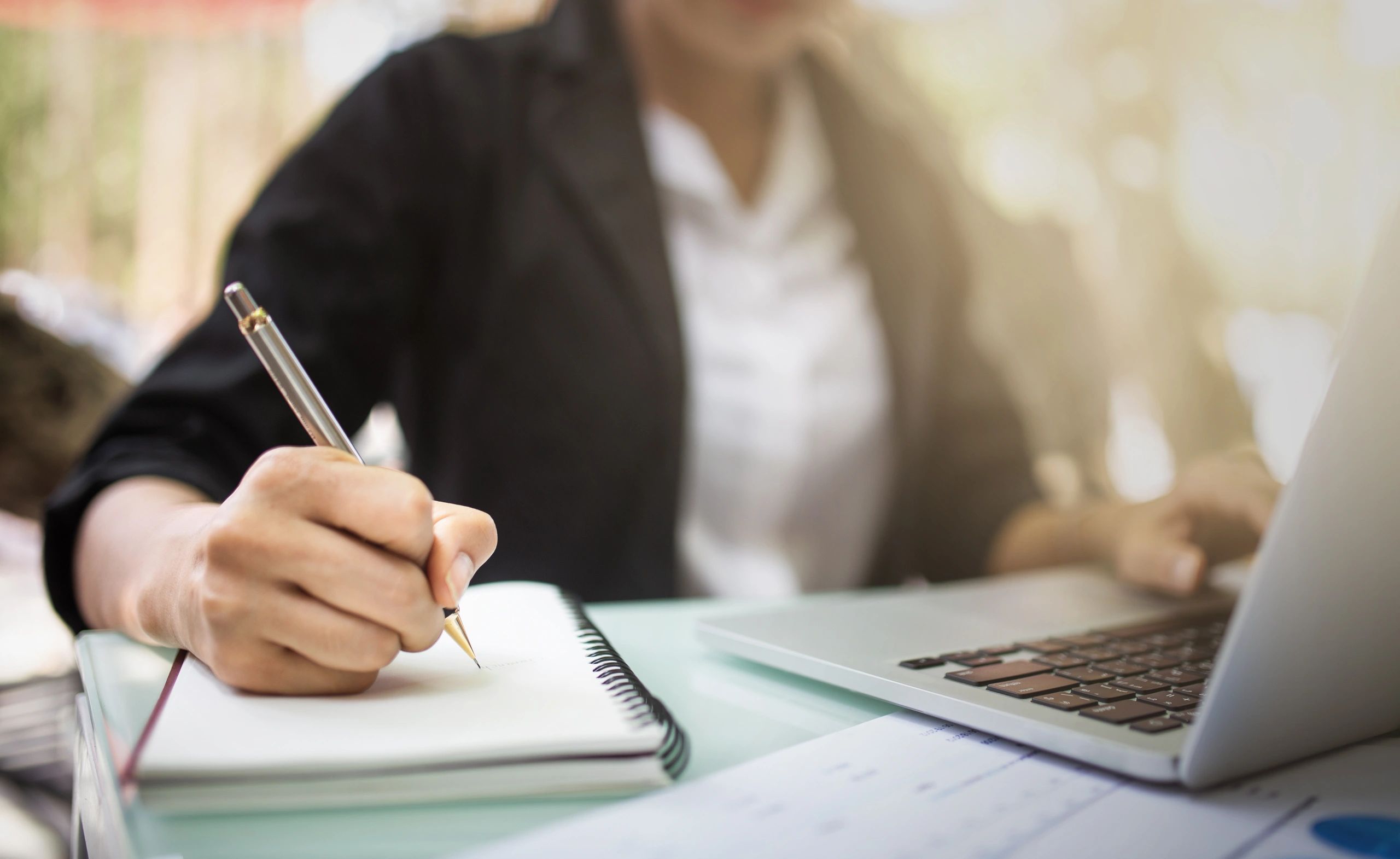 A person writing on paper with pen and laptop